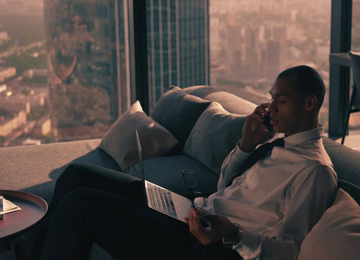 Executive man sitting on couch in high-rise apartment talking on the phone and working on laptop