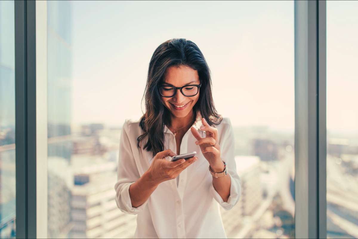 40 year old woman smiling at her phone and standing in front of window with city in background
