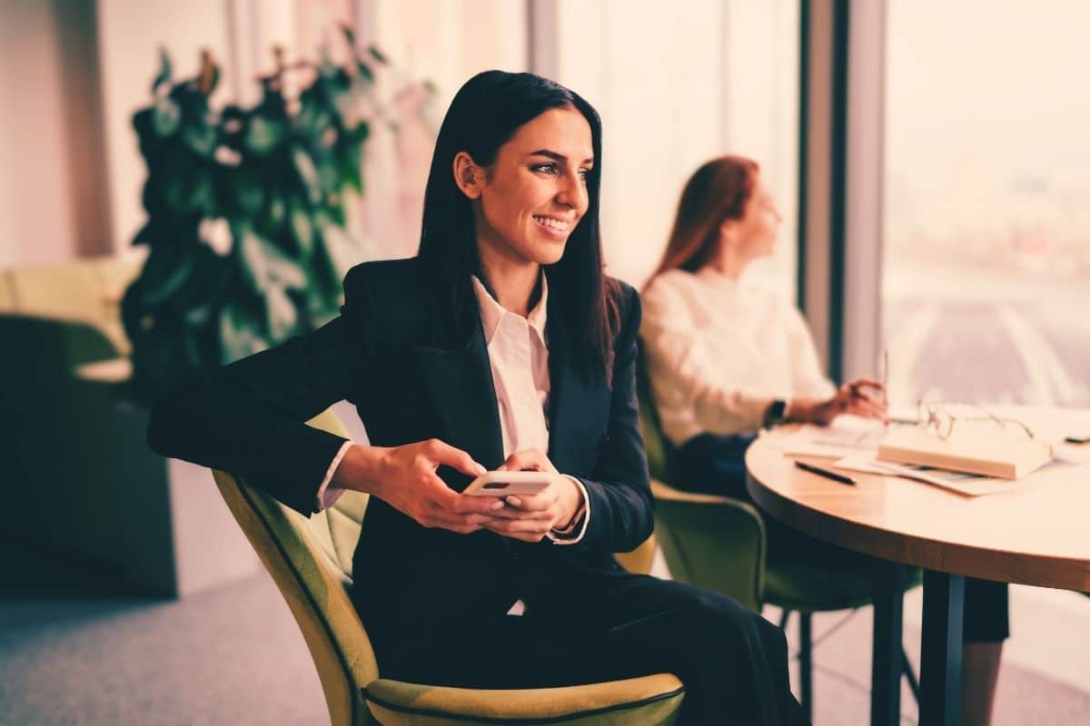 Business woman wearing a black suit and holding a cell phone sitting at a table inside an office suite