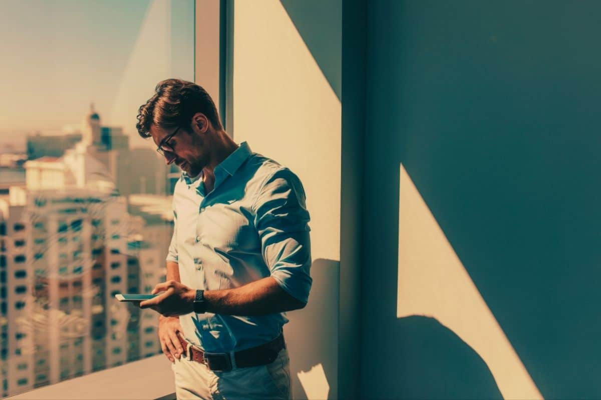 Business man looking at his cell phone and standing next to a window of a skyscraper